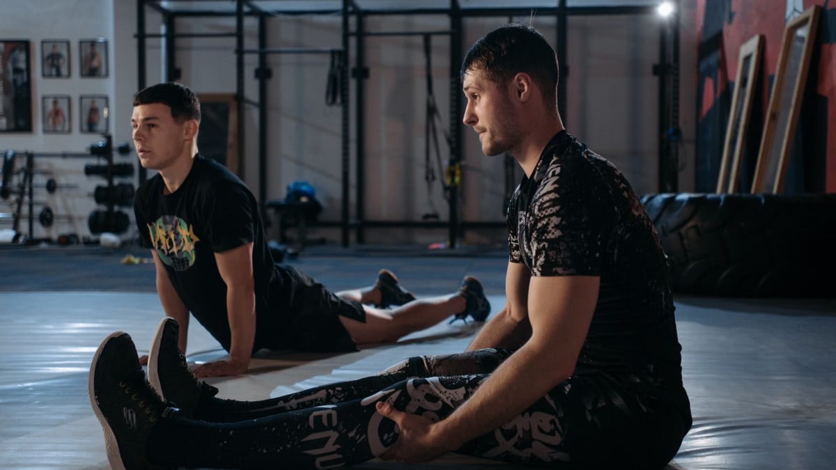 Two men performing MMA stretching exercises on a gym mat indoors.